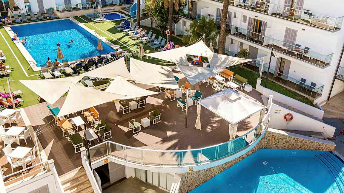 Et billede af et hotelresort med en stor pool omgivet af liggestole og parasoller. I forgrunden er en udendørs bar med mange siddepladser under skyggende sejl. (AI-genereret tekst)