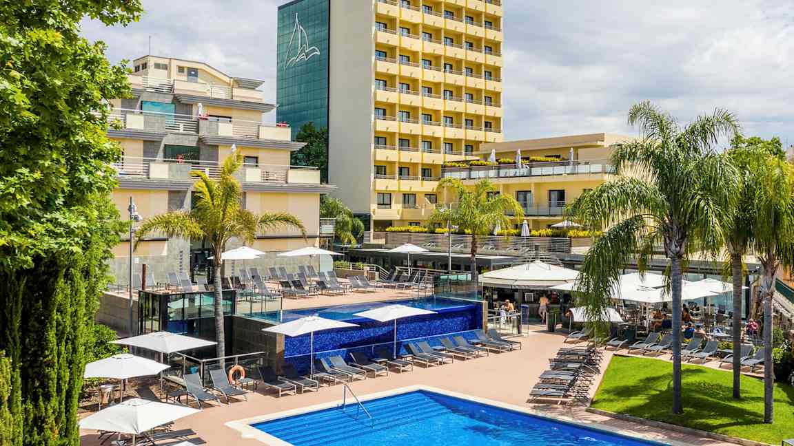 Billede af et hotel med gule facader, omgivet af palmer og en pool med liggestole på et soldæk. (AI-genereret tekst)