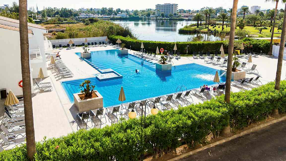 Udendørs swimmingpool med liggestole og parasoller omkring, omgivet af grønne planter og med udsigt over en sø og et åbent landskab. (AI-genereret tekst)