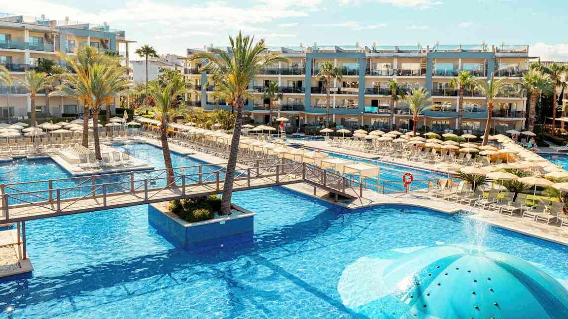 Poolområde ved et hotel med liggestole og parasoller, omgivet af palmer og bygninger. (AI-genereret tekst)