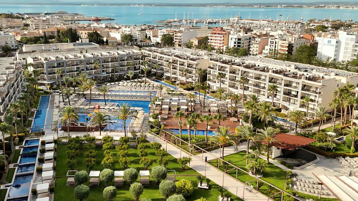 Et hotelkompleks med pools omgivet af grønne områder og liggestole, beliggende ved vandet med en havn i baggrunden. (AI-genereret tekst)