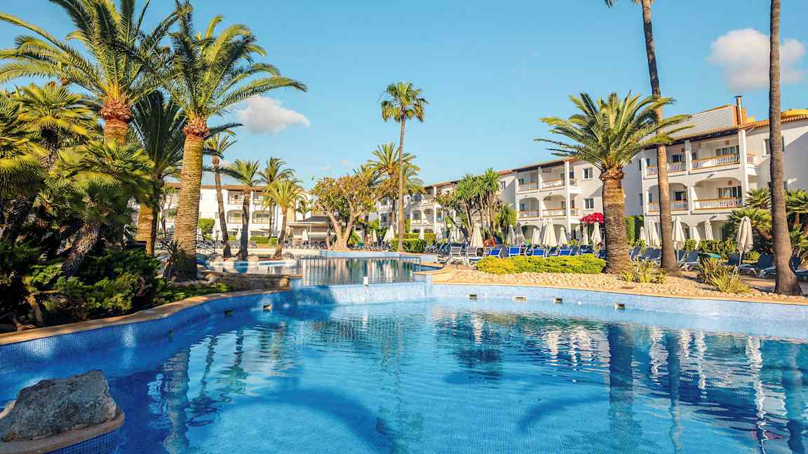 Udendørs pool med palmer og hvidmalet hotelbygning i baggrunden. (AI-genereret tekst)