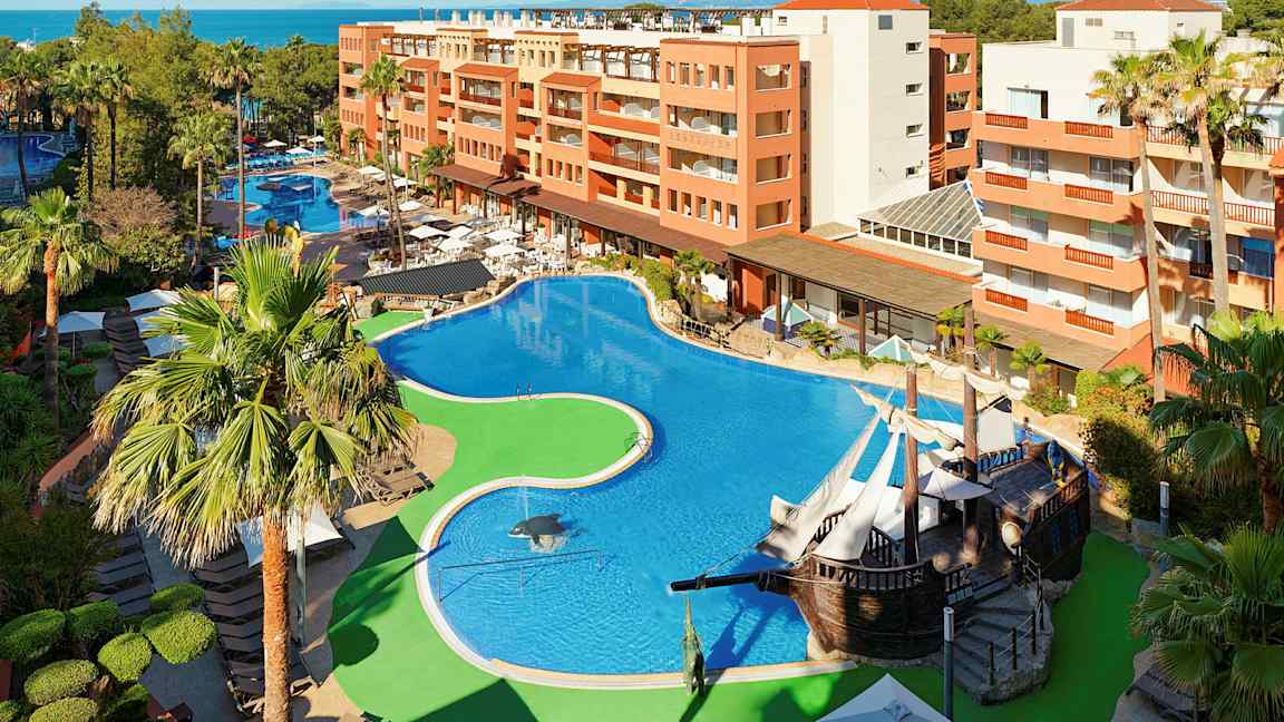 Hotelområde med stor pool, omgivet af palmer og bygninger. (AI-genereret tekst)