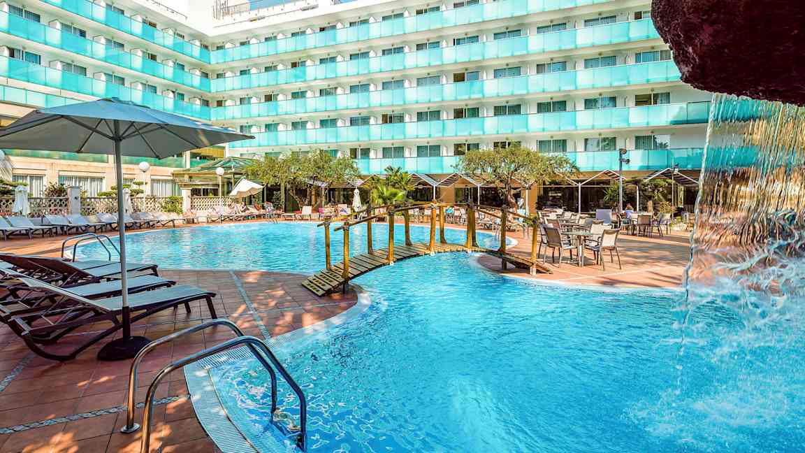 En udendørs swimmingpool ved en hotelbygning med liggestole, borde og parasoller. En lille bro over poolen og en del af en vandfaldsstruktur ses. (AI-genereret tekst)