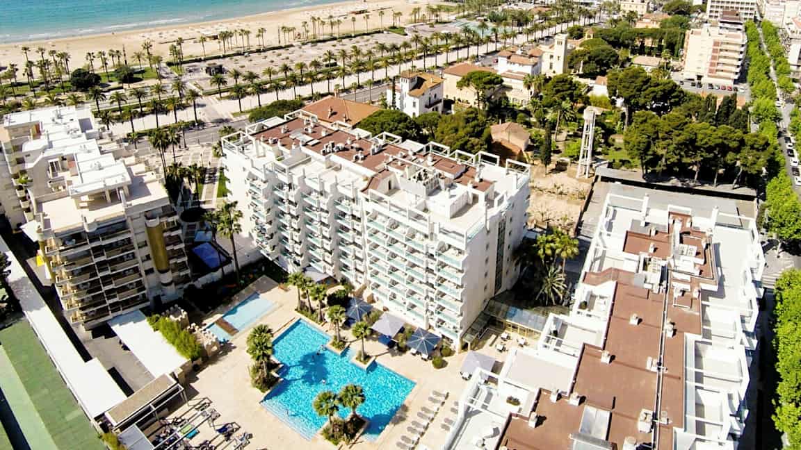 Luftfoto af et hotel med poolområde, omgivet af bygninger og grønne områder. I baggrunden ses en sandstrand og havet. (AI-genereret tekst)