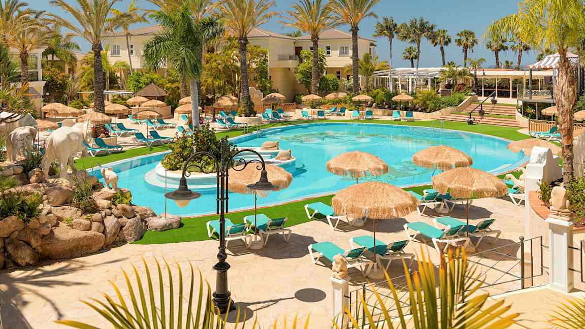 Et billede af et luksuriøst feriested med en pool, omgivet af liggestole og parasoller, omringet af palmer og bygninger. (AI-genereret tekst)