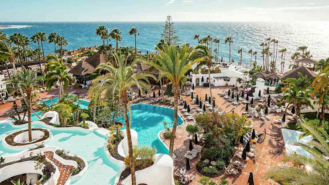 Udsigt over et hotelområde med to pooler, palmer og solsenge ved havet. (AI-genereret tekst)