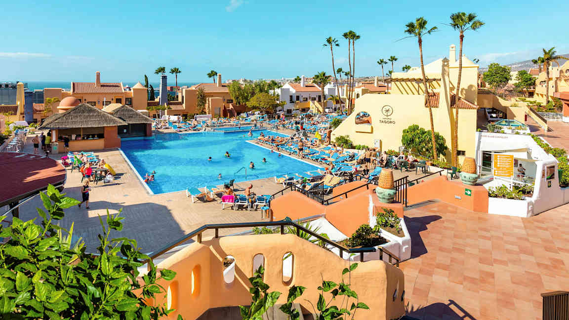 Et hotelområde med en stor pool omgivet af liggestole og bygninger. (AI-genereret tekst)