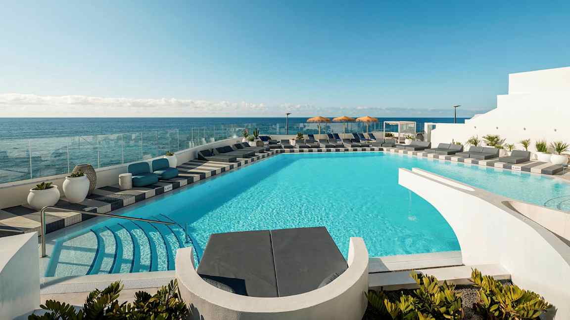 En pool med turkis vand omgivet af solsenge og udsigt over havet. I baggrunden ses en klar blå himmel. (AI-genereret tekst)