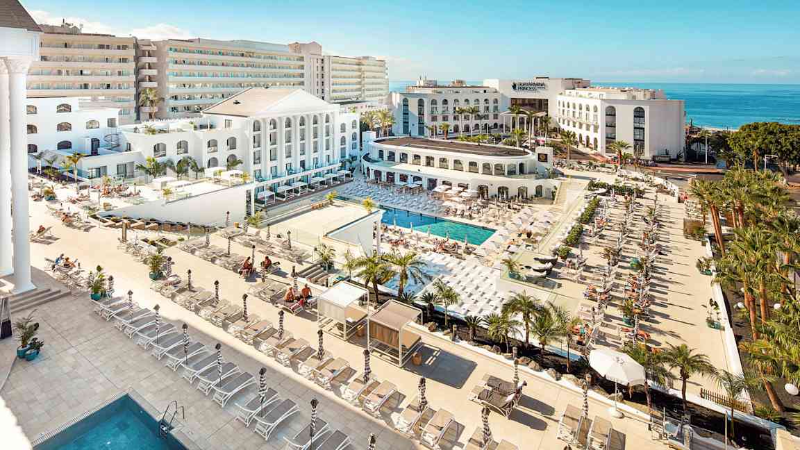 Udsigt over et hotelområde med pools, solsenge og hvid arkitektur tæt på havet. (AI-genereret tekst)