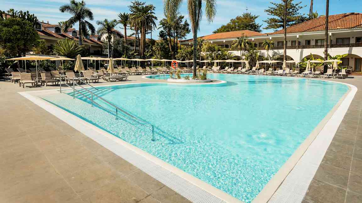 En udendørs pool omgivet af liggestole og parasoller med palmer i baggrunden. (AI-genereret tekst)