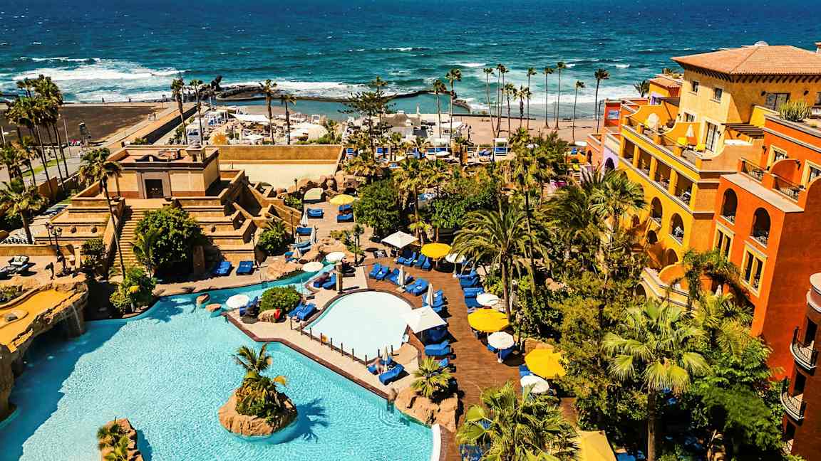 Et hotelområde med pools, beplantning og farverige bygninger ved havet. (AI-genereret tekst)