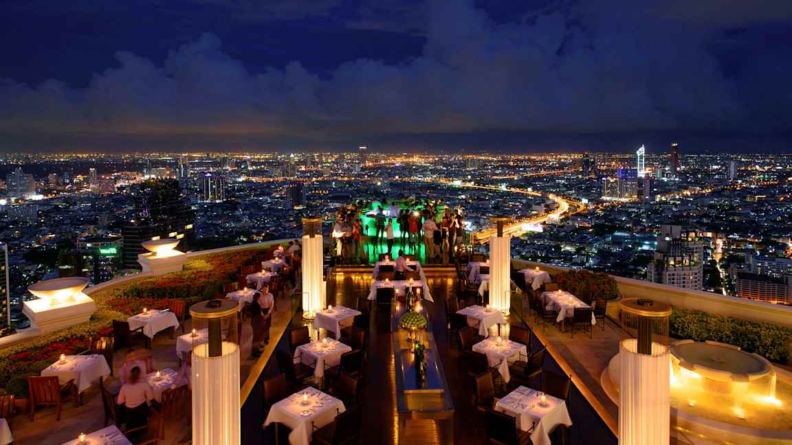 En tagterrasse-restaurant om aftenen med oplyste borde og udsigt over en by med lysende lys. (AI-genereret tekst)