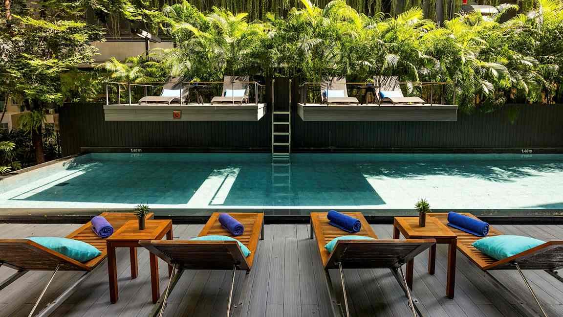 En pool med liggestole og borde foran, omgivet af grøn beplantning og palmer. (AI-genereret tekst)