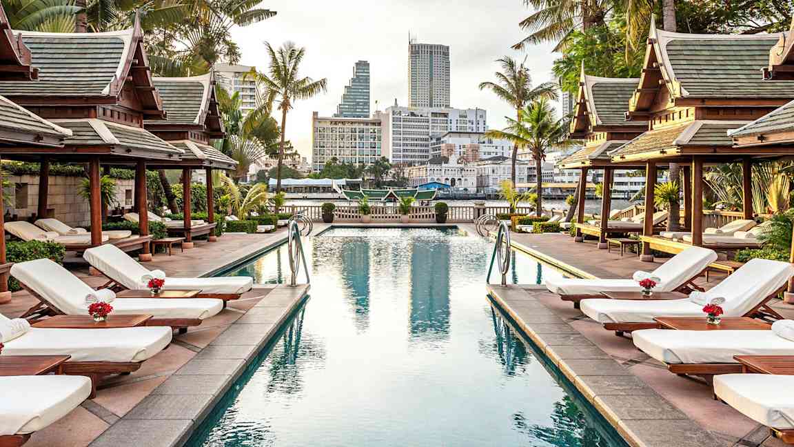 Et poolområde omgivet af liggestole, palmer og pavilloner i traditionel stil med en by i baggrunden. (AI-genereret tekst)