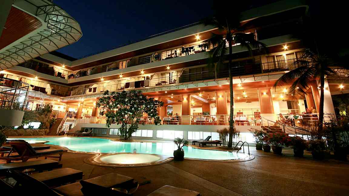 Aftenudsigt af en fleretagers bygning med oplyst facade og en pool i forgrunden. (AI-genereret tekst)