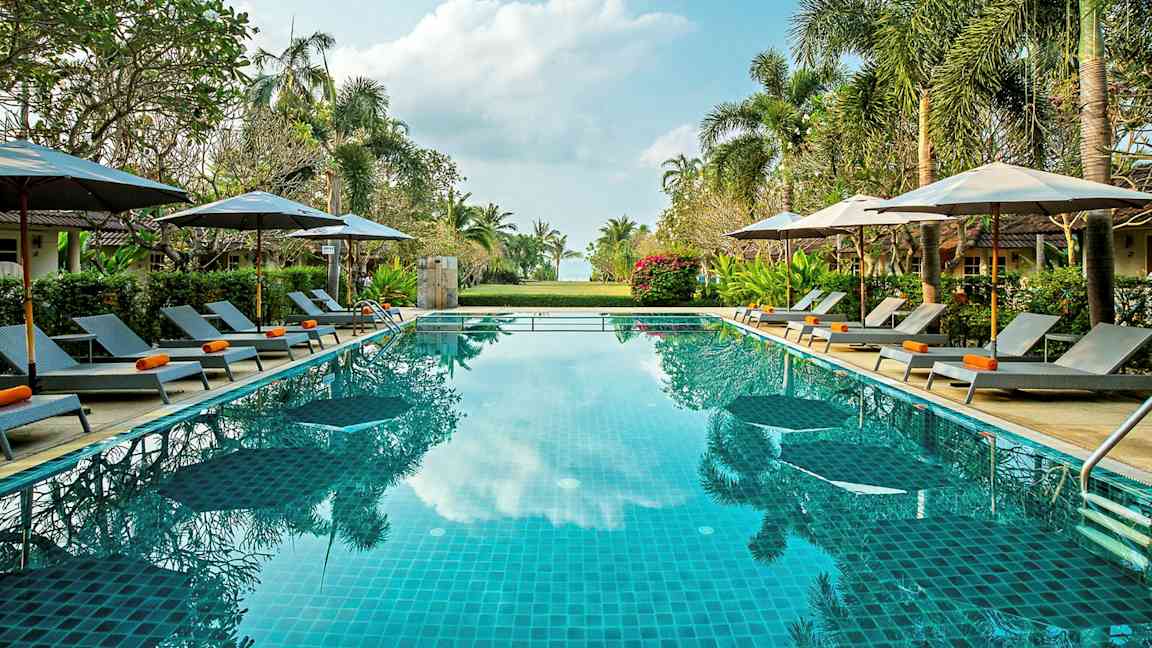 Billede af en udendørs swimmingpool omgivet af liggestole, parasoller og grønt under en klar himmel. (AI-genereret tekst)