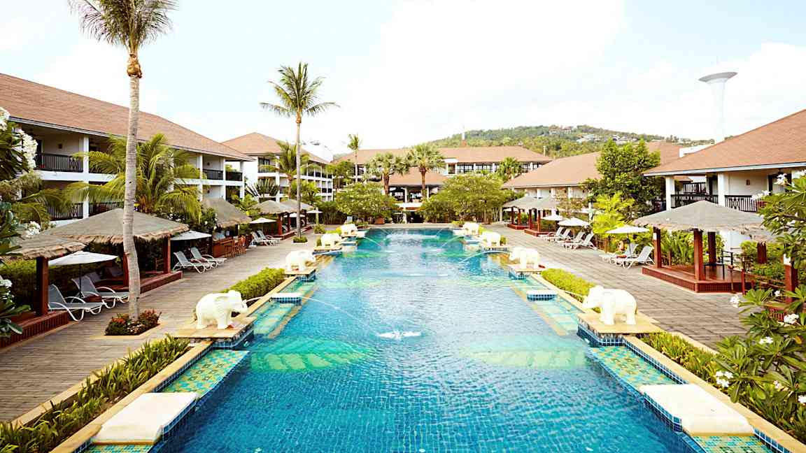 En stor pool på et feriested omgivet af palmer, liggestole og bungalowlignende bygninger. (AI-genereret tekst)