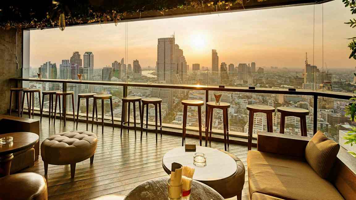 En moderne indendørs bar med høje barstole og borde foran et stort vindue med udsigt over en bysilhuet ved solnedgang. (AI-genereret tekst)