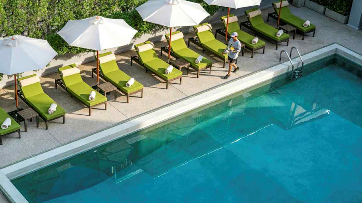 Pool med liggestole og parasoller ved kanten, samt en person der går forbi. (AI-genereret tekst)