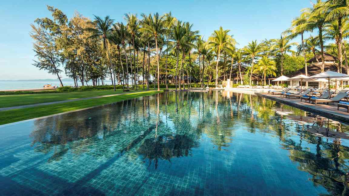 En udendørs swimmingpool omgivet af grønne områder og palmer, med udsigt over stranden og havet. (AI-genereret tekst)