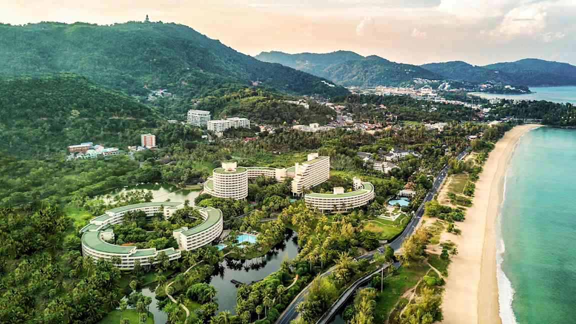 Luftfoto af et kystområde med hotelbygninger, omgivelser med grønt og en strand. (AI-genereret tekst)