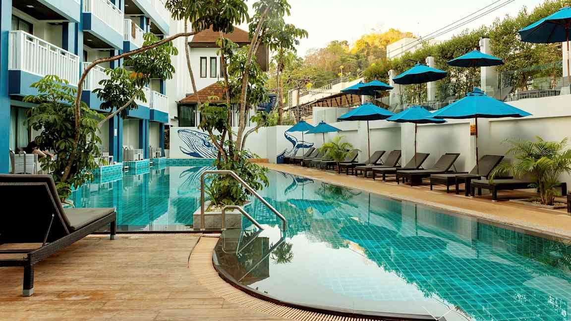 En udendørs swimmingpool med liggestole og parasoller, omgivet af vegetation og en fler-etagers hotelbygning. (AI-genereret tekst)