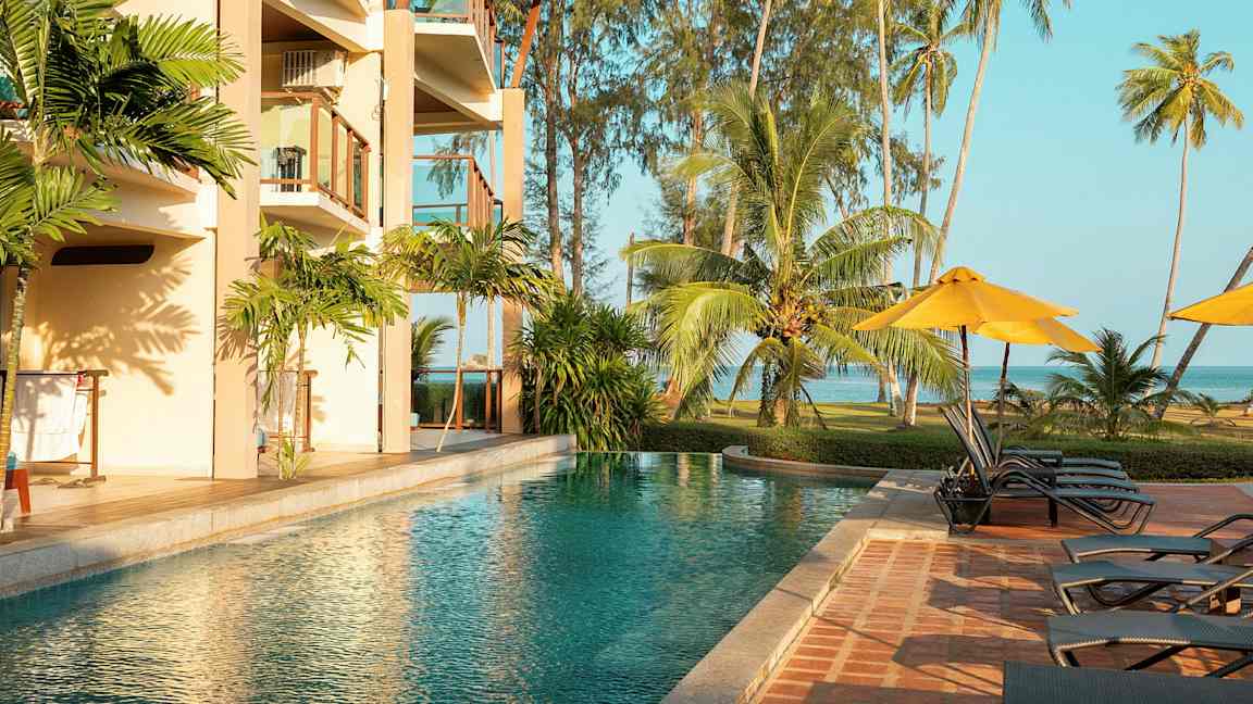 En udendørs pool ved siden af et hotel med altaner, omgivet af tropiske palmer og græsareal ved havet. (AI-genereret tekst)