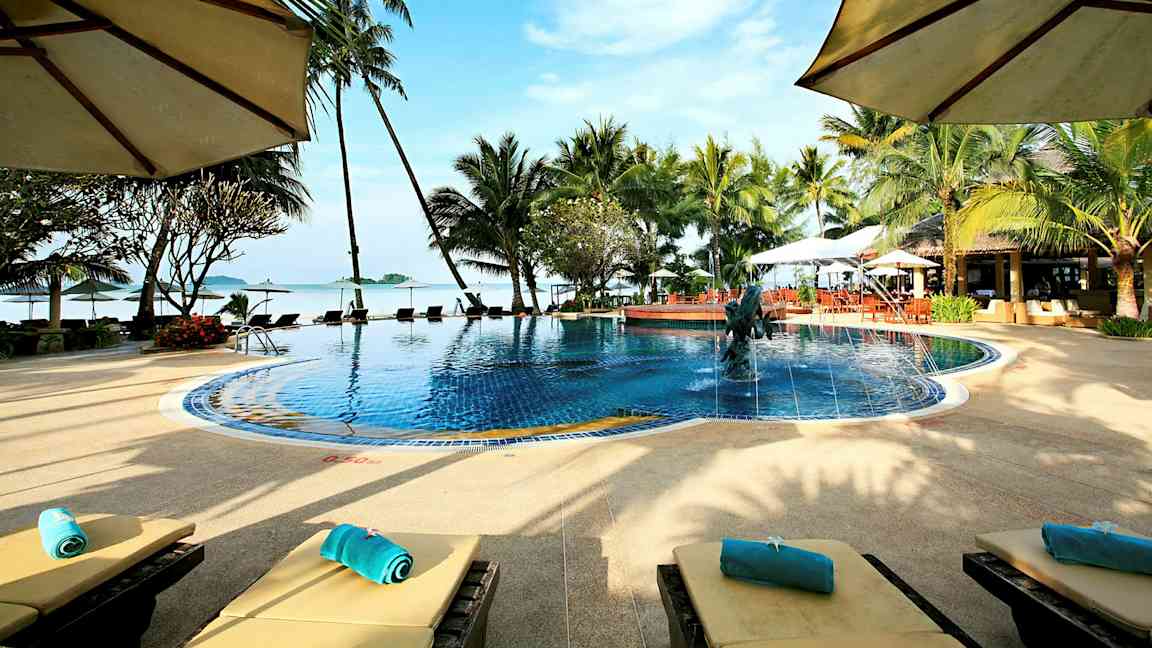 En pool omgivet af palmer og liggestole med udsigt over havet og en blå himmel. (AI-genereret tekst)