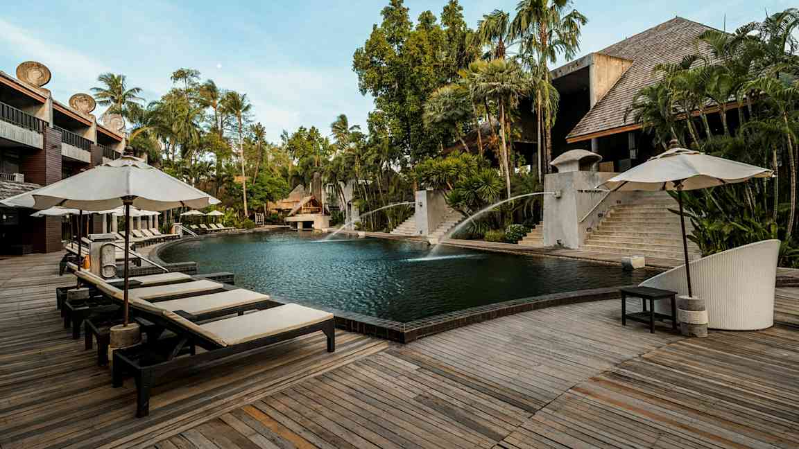 En udendørs pool omgivet af trædæk og liggestole med parasoller. Der er frodig beplantning og bygninger af træ og sten i baggrunden. (AI-genereret tekst)