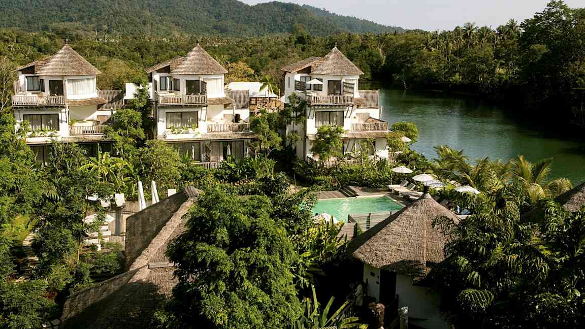 Et hotelområde med huse i tropisk stil, omgivet af grønt og en flod i baggrunden. (AI-genereret tekst)
