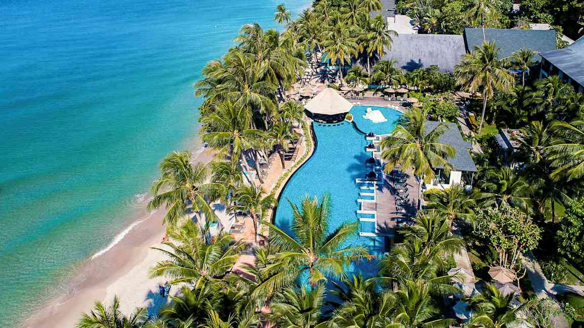 Luftfoto af en strandresort med pool, liggestole og palmer nær havet. (AI-genereret tekst)