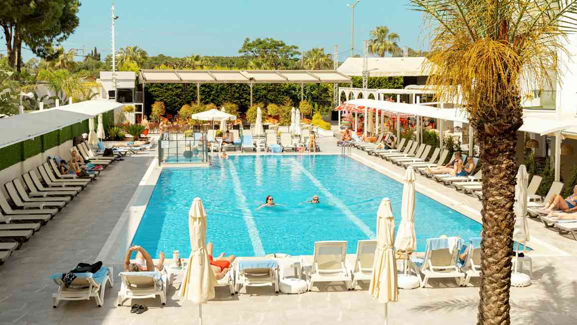 Billede af en pool omgivet af liggestole og parasoller, med personer der svømmer og soler sig. (AI-genereret tekst)