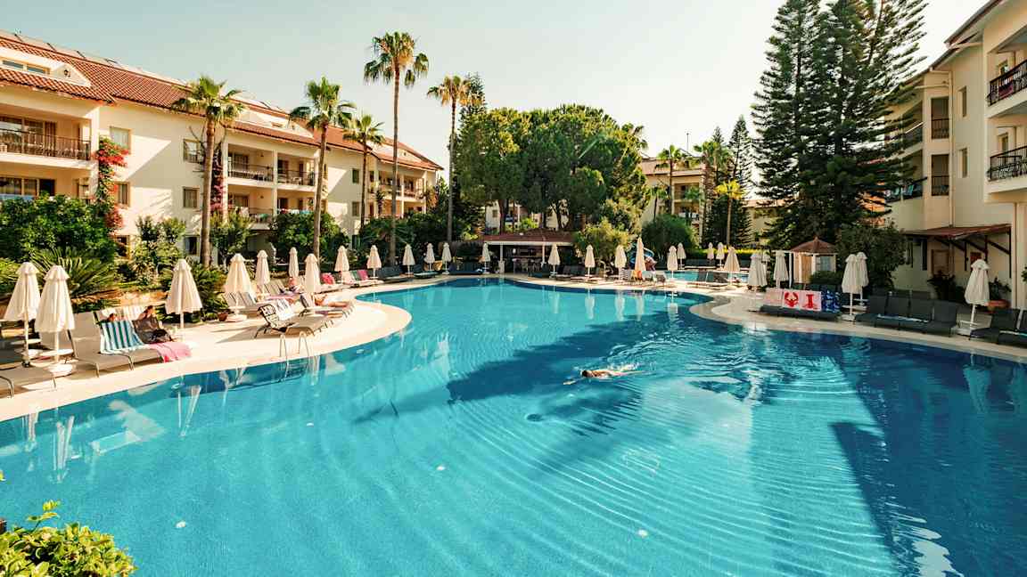 Stor udendørs swimmingpool omgivet af liggestole og parasoller, med grønne træer og hotelbygninger i baggrunden. (AI-genereret tekst)