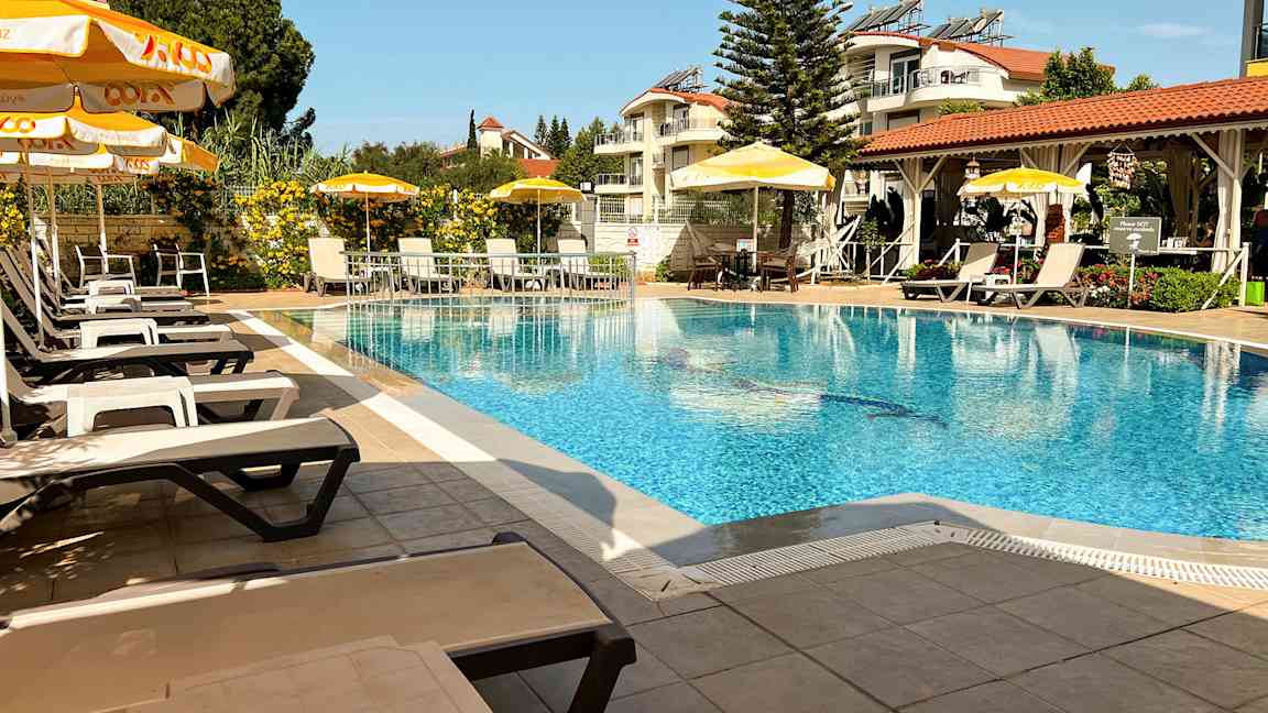 En udendørs pool med liggestole og parasoller, omgivet af grønne områder og bygninger. (AI-genereret tekst)