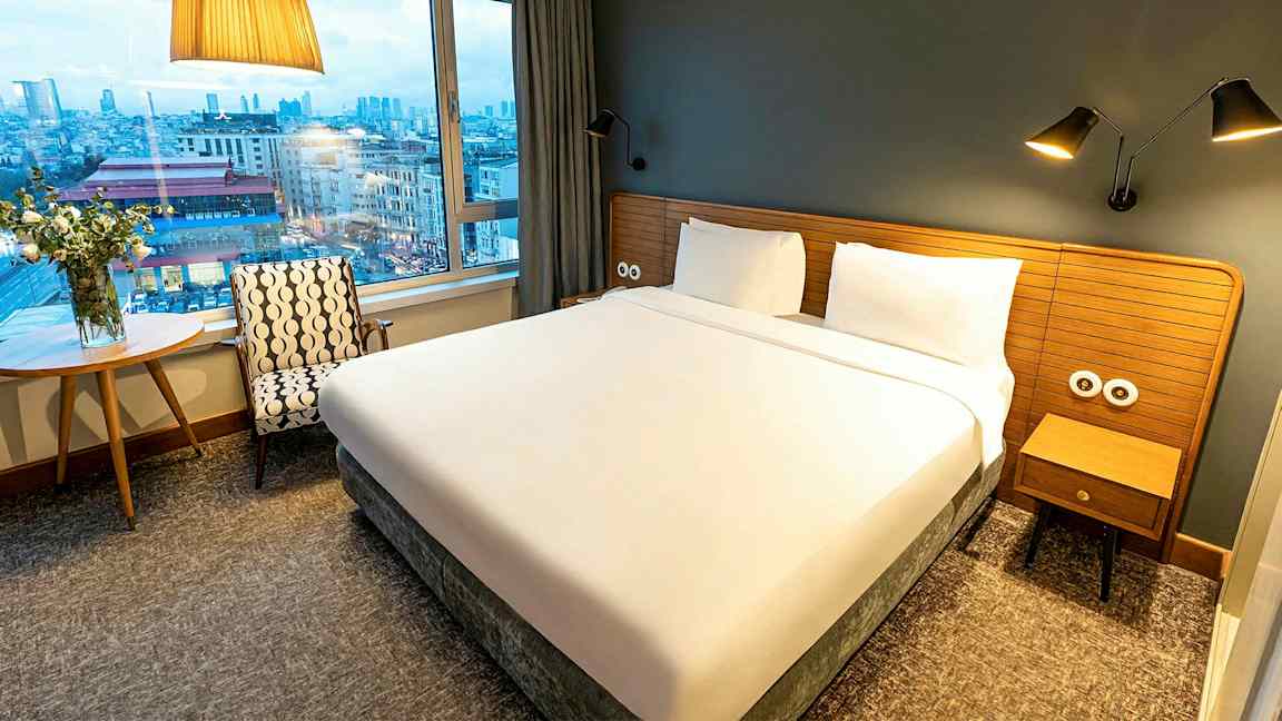 Et hotelværelse med en stor seng, vindue og udsigt over en by. (AI-genereret tekst)