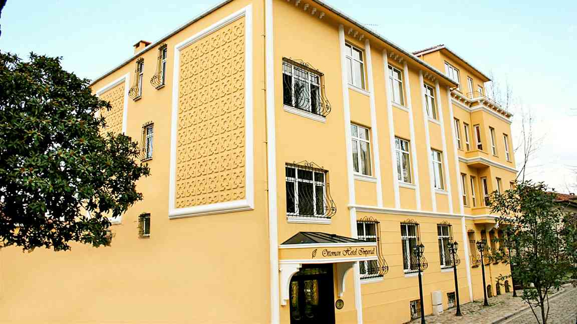 Et gult hotel med dekorativ facade, vinduer med jerngitre og en indgang med overdækning. (AI-genereret tekst)