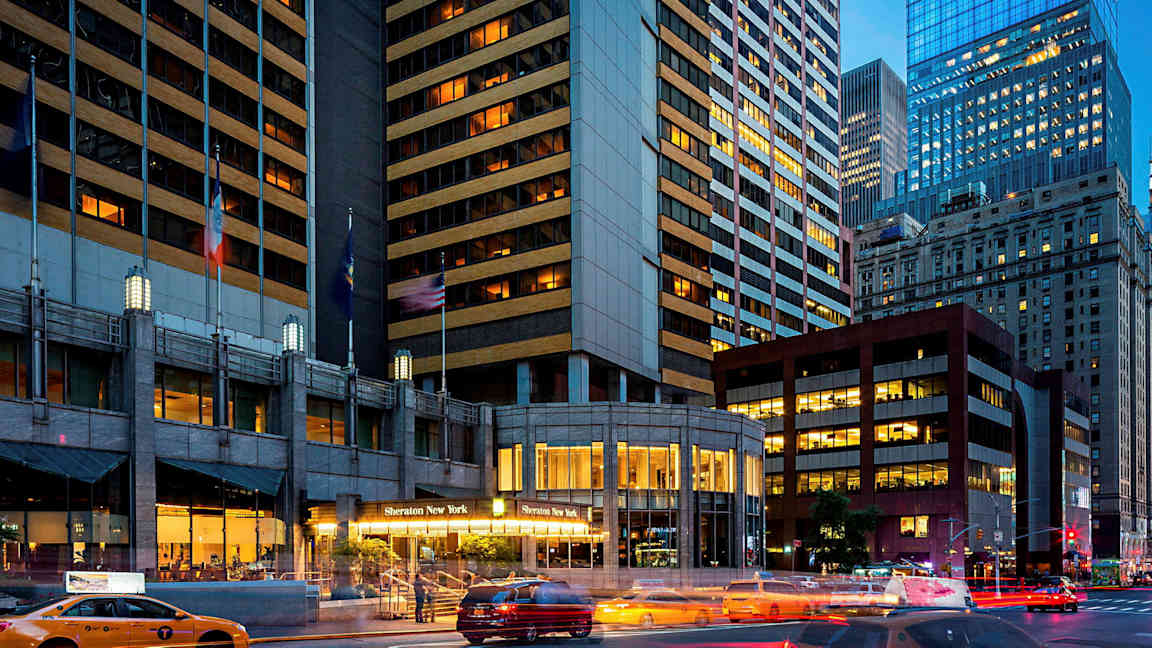 Et billede af et bymiljø med høje bygninger, hvoraf en bygning har teksten "Sheraton New York" ved indgangen. Taxaer er synlige på gaden. (AI-genereret tekst)