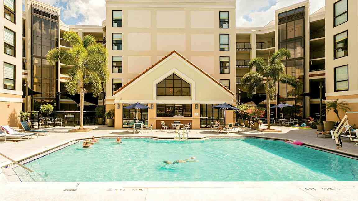 Billede af et hotel med en swimmingpool i forgrunden. Personer svømmer og slapper af ved poolområdet. Hotellets facade ses i baggrunden med palmer i nærheden. (AI-genereret tekst)