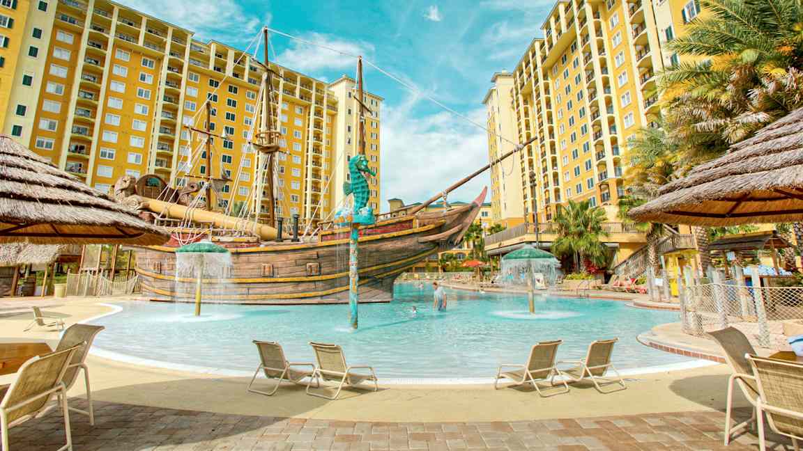 En pool omgivet af liggestole og gule lejlighedsbygninger med et piratskib i midten. (AI-genereret tekst)
