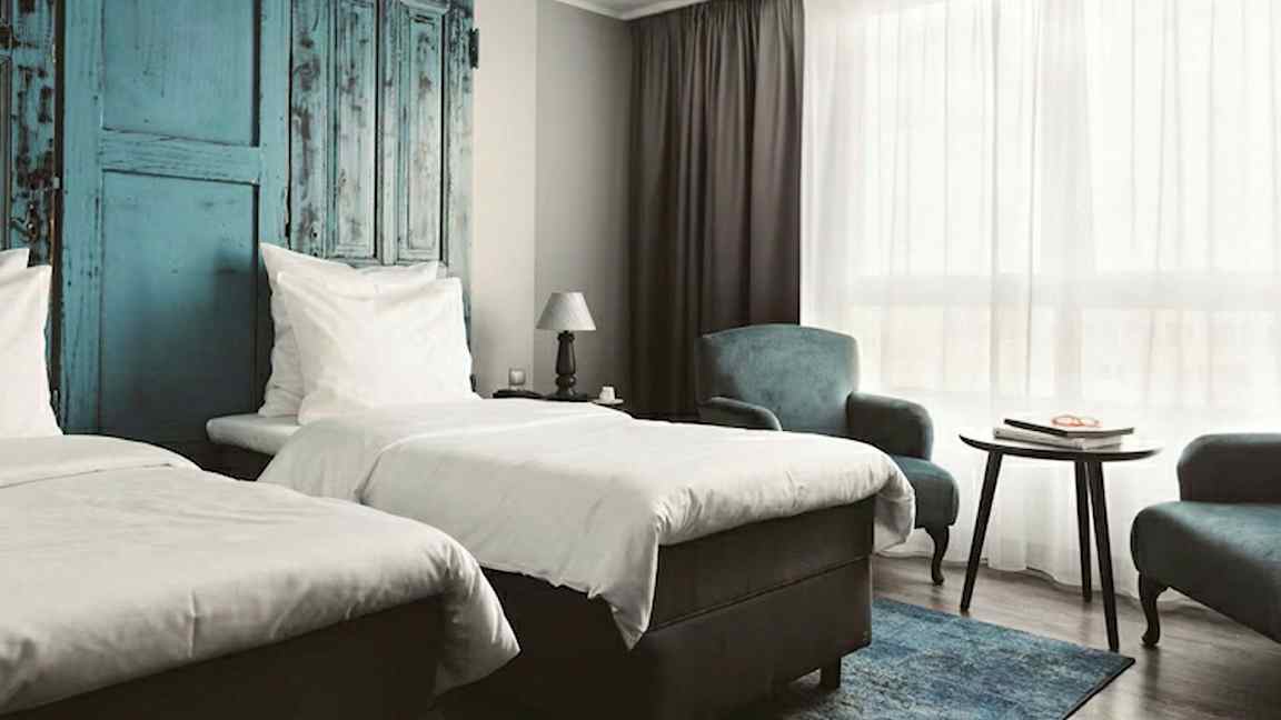 Et hotelværelse med to enkeltsenge, blå detaljer på væggen, et bord og lænestole samt et stort vindue med hvidt gardin. (AI-genereret tekst)