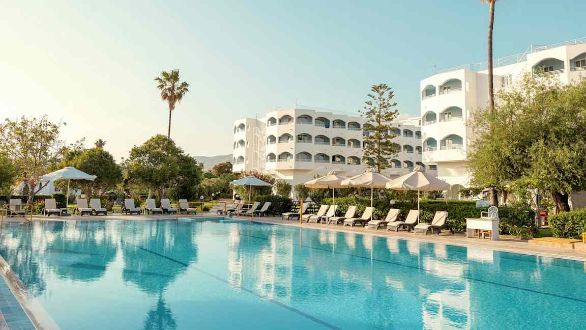 Hotelbygning med pool, liggestole og parasoller i forgrunden. (AI-genereret tekst)