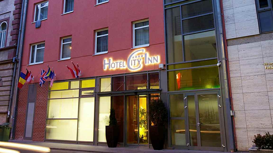 Uden for en hotelbygning med et skilt, der lyder "Hotel City Inn", og flere flag ved indgangen. (AI-genereret tekst)