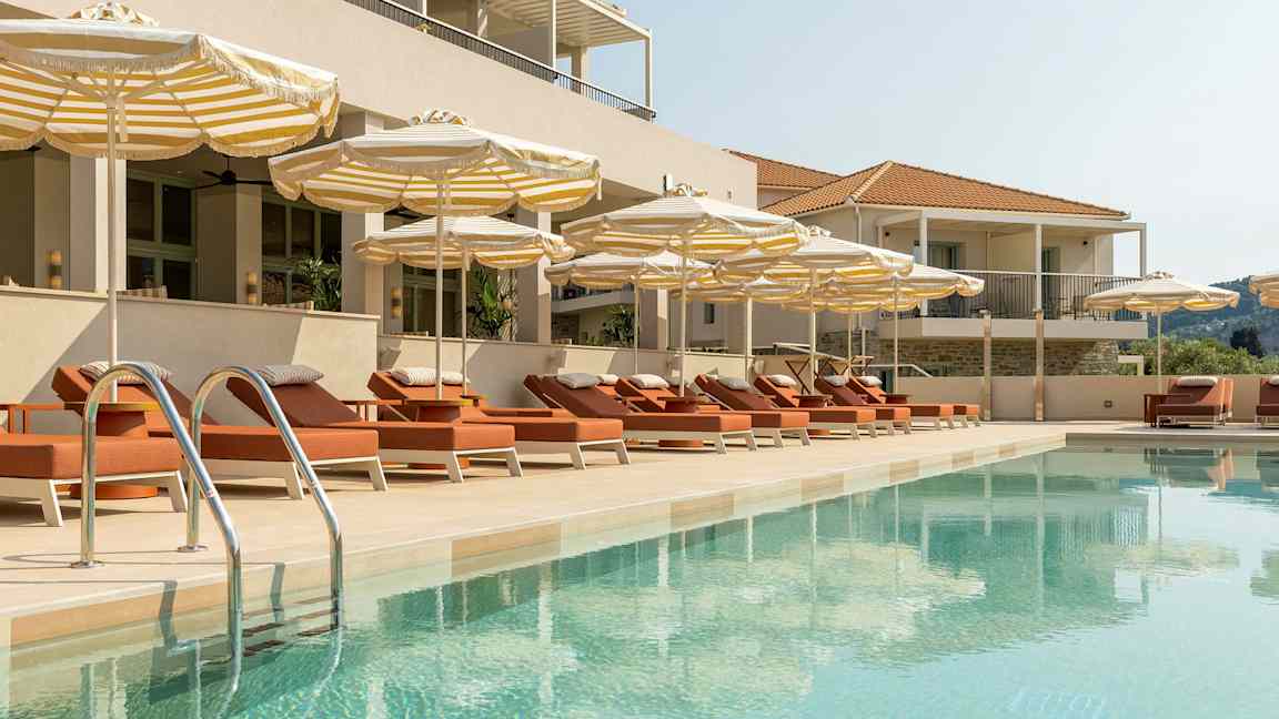 En swimmingpool omgivet af liggestole og parasoller ved en bygning. (AI-genereret tekst)