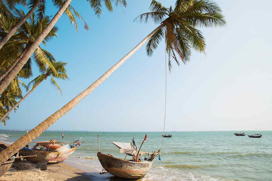 Udsigt over en tropisk strand med palmer og traditionelle både ved vandet. (AI-genereret tekst)
