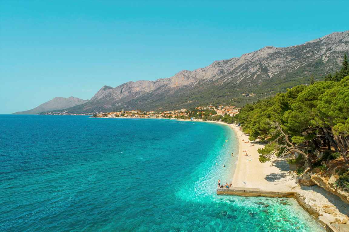 Udsigt over en stenstrand med turkis vand og frodige træer. Bjerge og en landsby ses i baggrunden. (AI-genereret tekst)