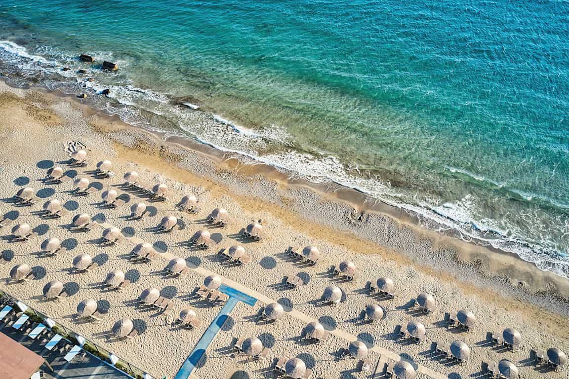 Luftfoto af en sandstrand med liggestole og parasoller, med kystlinjen og blåt hav i baggrunden. (AI-genereret tekst)