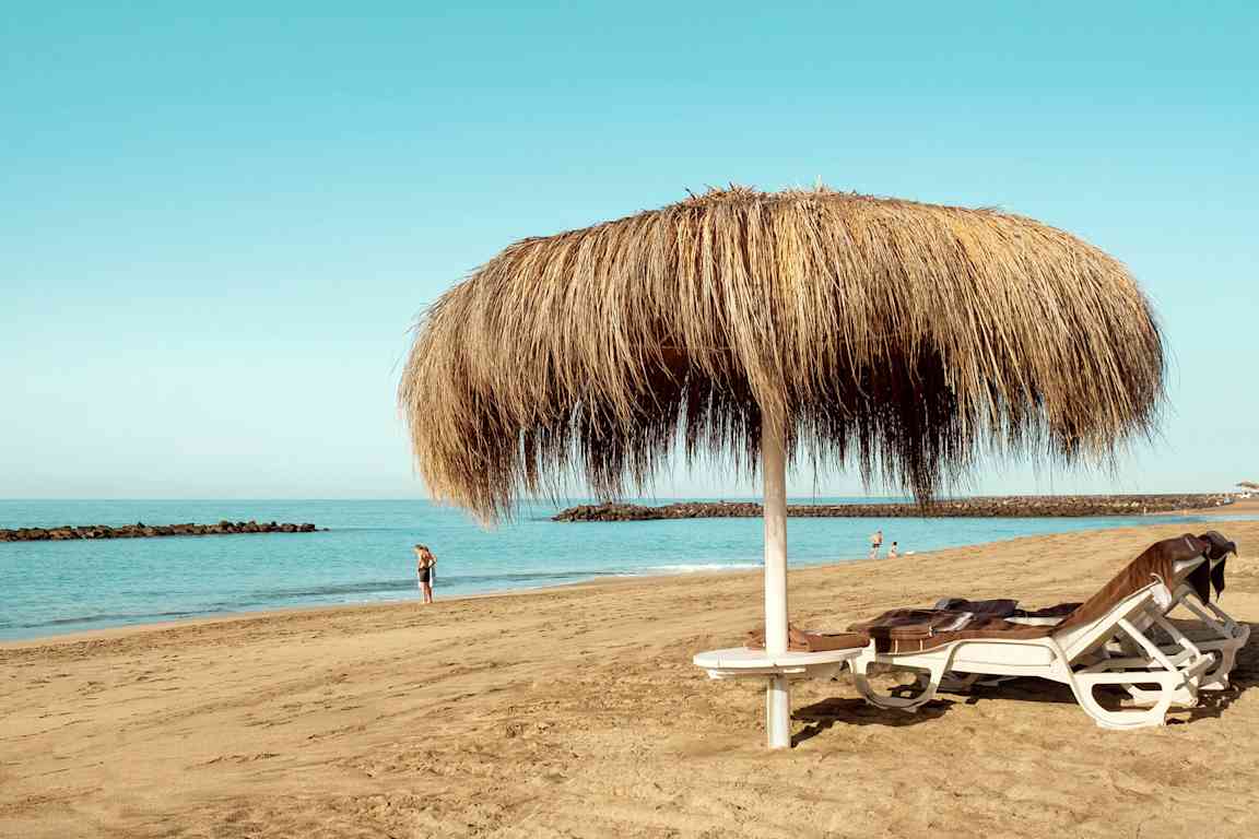 Billede af en sandstrand med to liggestole under en halmparasol samt personer, der går langs vandet. (AI-genereret tekst)