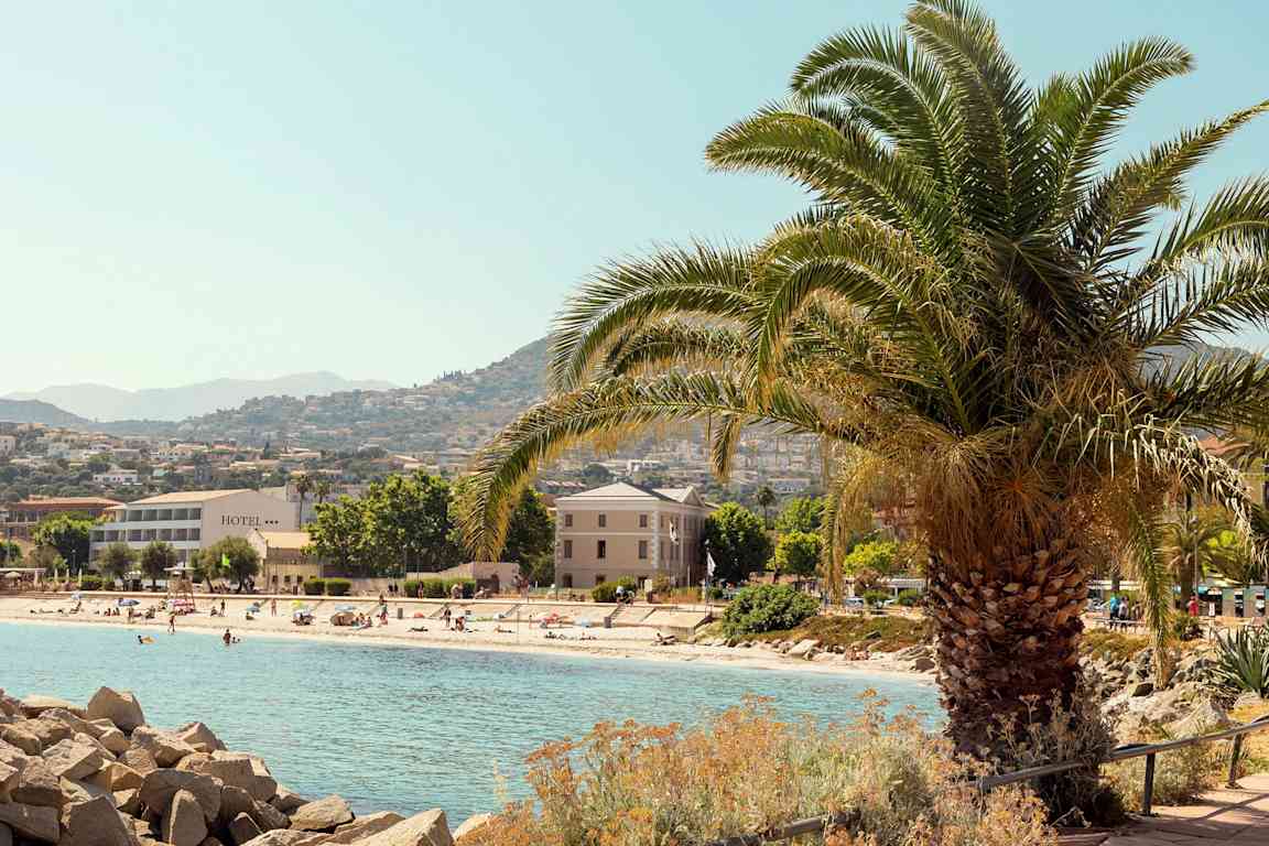 En strand med badegæster, omgivet af hotelbygninger og træer med bjerge i baggrunden. En stor palme står i forgrunden. (AI-genereret tekst)