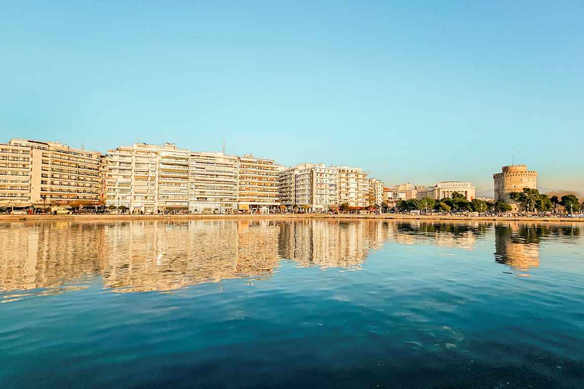 En by ved vandet med bygninger og et rundt tårn reflekteret i det rolige vand. (AI-genereret tekst)